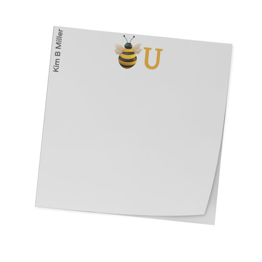 A white 3x3 inch sticky notes with a bee illustration and the letter 'U' on them, designed by Kim B Miller, angled view.